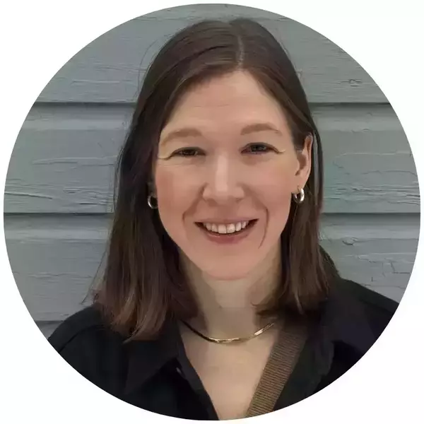 Sarah Hill's headshot. A smiling woman with brown hair, wearing silver hoop earrings, a gold necklace, and a dark shirt, against a light blue wooden wall.