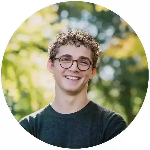 Finnbarr Munley's headshot. Young man with curly brown hair, tortoiseshell glasses, and a dark green sweater smiling broadly against a blurry green outdoor background.